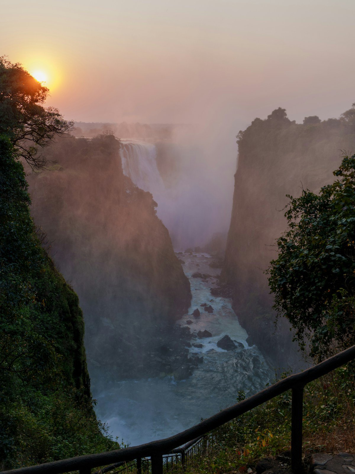 Victoria Falls gorge at sunrise with mist rising from the Zambezi River below and lush green vegetation on the cliff edges